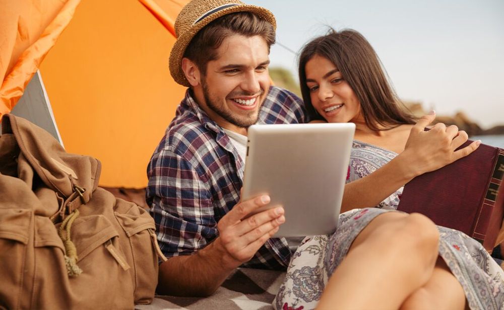 Couple sitting at tent using tablet computer and reading book